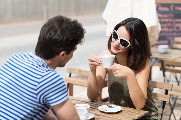 Young-woman-in-stylish-sunglasses-flirting-with-a-man-over-coffee-at-an-open-air-cafeteria-smiling-at-him-with-a-teasing-look